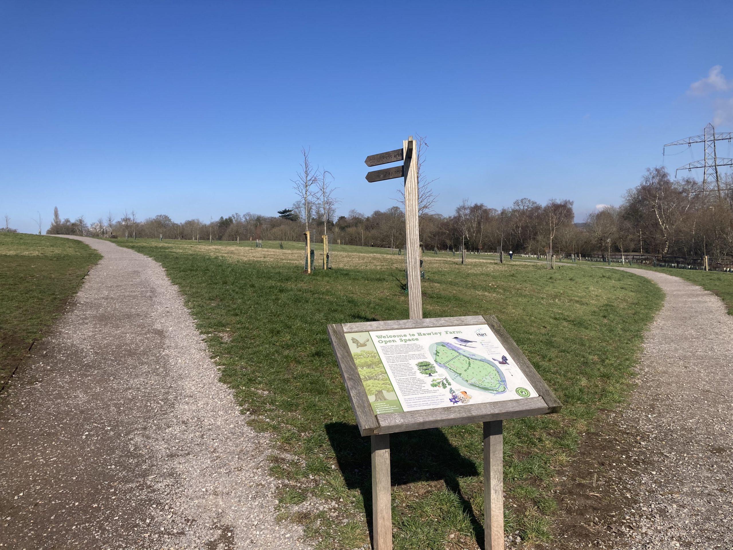 Photo of an open grass area with gravel paths, a large waymarker post and an interpretation panel. In winter blue sky.