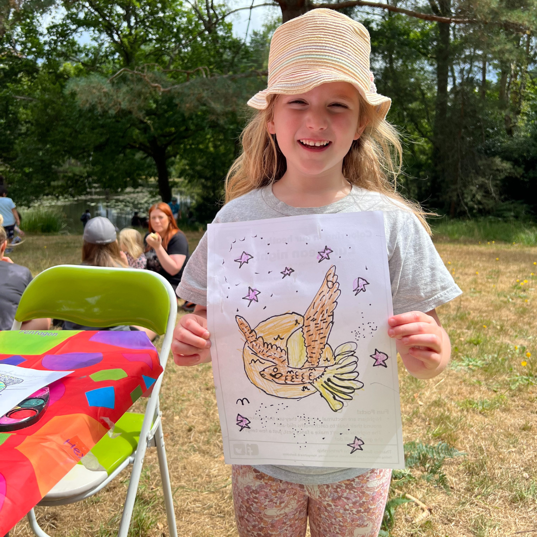 Photo of a little girl in the sunshine, holding up a picture she drew of a nightjar.