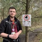 Photo of a man standing beside a sign tied to a tree. The sign asks that we keep to main paths.