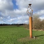 Photo of a tall metal sculpture. Curving rods carry a skylark high into the sky.