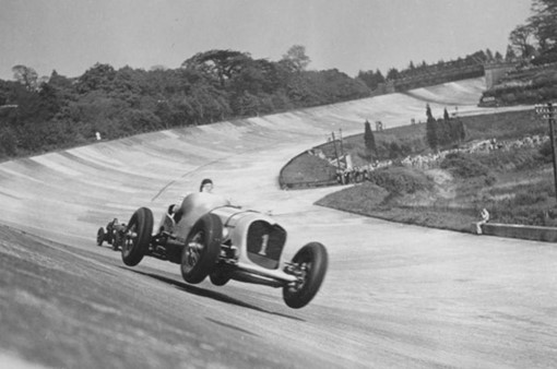 Old black & white photo of an old fashioned sports car hurtling around a pristine banked track.