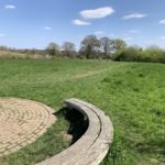 A photo with a curved bench in the foreground and people walking a dog in the distance.