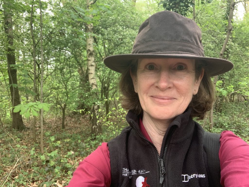 Photo of Sarah in a brimmed hat, with leafy woodland scene behind her.