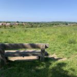 Photo of a wooden bench looking out at the view across meadows and nearby built up areas.