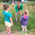 A ranger in a green top holds out a pot for two small girls to look at. I think the pot contains an insect.