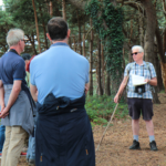 Photo of a man in sunglasses, holding notes and a walking stick. He's talking to a group of people.