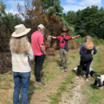 Photo of a group of people on a heathland path. The vegetation is burnt to one side. Warden Henry stands with his arms outstretched, obviously explaining something to the attentive group.