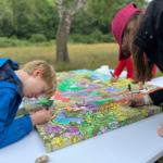 Photo of two people bent over a paiting, working on the colouring in of a couple of section. They look very concentrated.