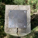 Photo of a brass rubbing plaque mounted on top of a post. The image shows an Adder.