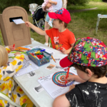 A small boy in a bright red hat posts a message to nature into a wooden postbox. A boy in the foreground wearing a colourful hat, is colouring in an Adder with bright colours.