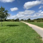 Photo of a grassy area with scattered trees. A gravel path leads up to a gateway.