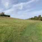 Photo of the slopes of a green knoll with people at the top enjoying the view.