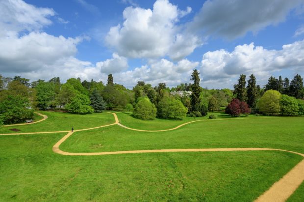 Photograph of a grassland park, criss crossed with surfaced paths. Trees flank the far slopes and large mansion house is just visible.