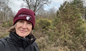 Selfie photo of Sarah in her burgundy "Thames Basin Heaths" woolly hat and heathland behind.