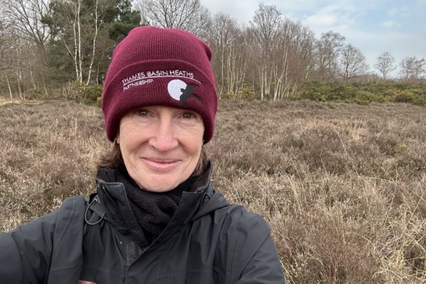 Selfie photo of Sarah in her burgundy "Thames Basin Heaths" woolly hat and heathland behind.