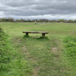 Photo looking out over a green meadow, a bench in the foreground. In the distance4 lots of people are using the bike track and sports areas.