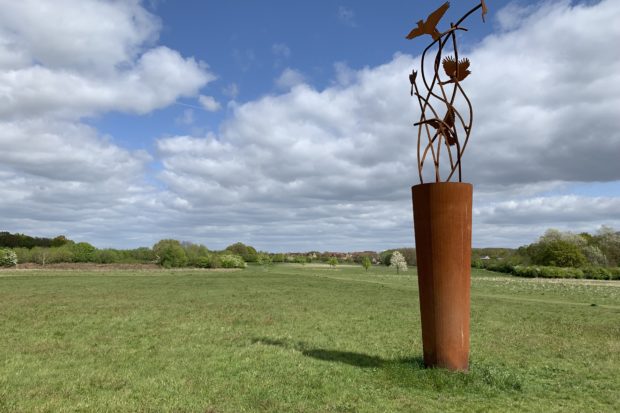 Photo of a large green meadow, with a iron artwork in the foreground. The artwork celebrates the Skylarks that nest here.