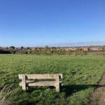 Photo of a viewpoint from a green meadow in autumn looking over a wooden bench towards housing